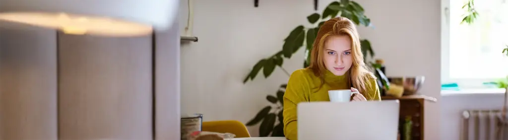 Junge Frau mit Kaffeetasse in der Hand blickt in ihren Laptop.