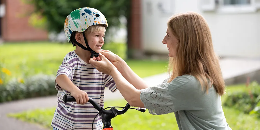 Eine Mutter zieht ihrem Kind einen Fahrradhelm an, da nicht nur finanzielle Kindervorsorge sinnvoll ist.